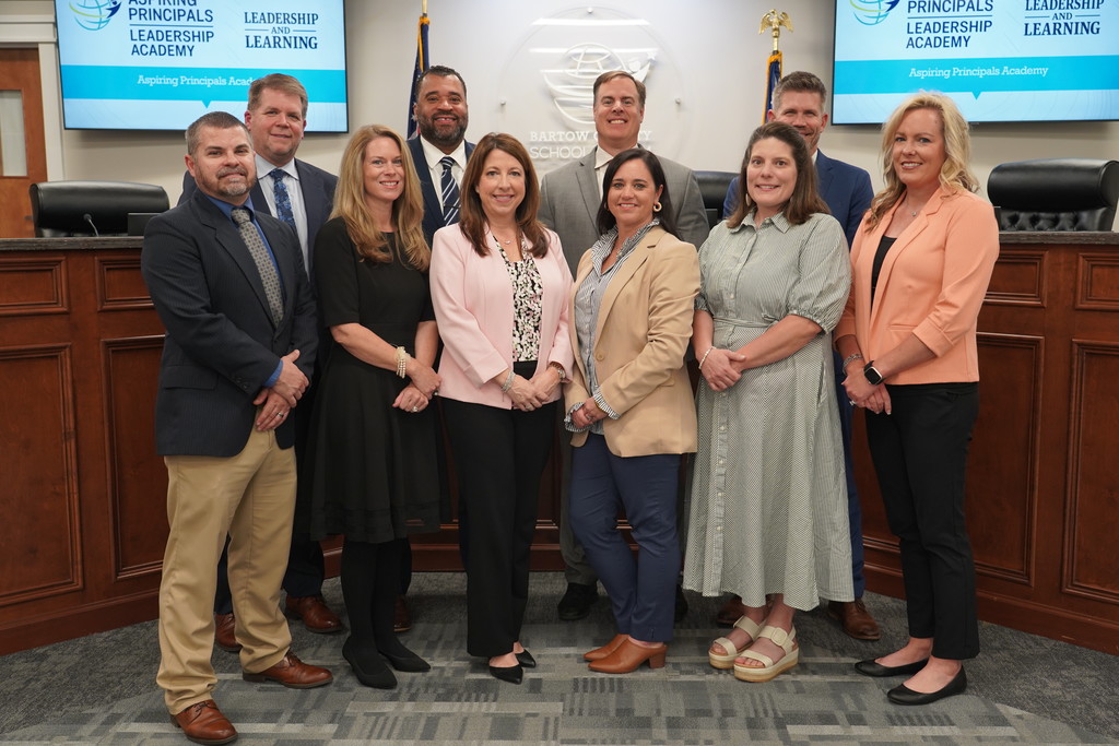 A group of adults, some in business attire, stand in a row inside a courtroom. A wooden bench is behind them.