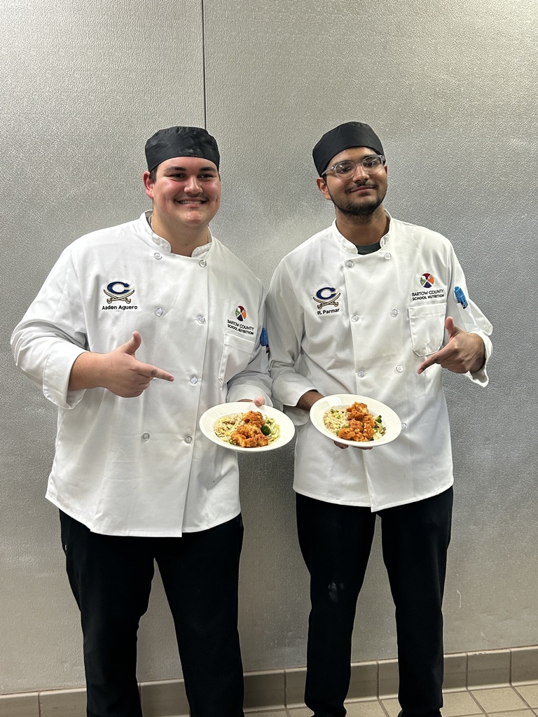 Two chefs in white coats, smiling, posing with plates of food in front of a silver wall.