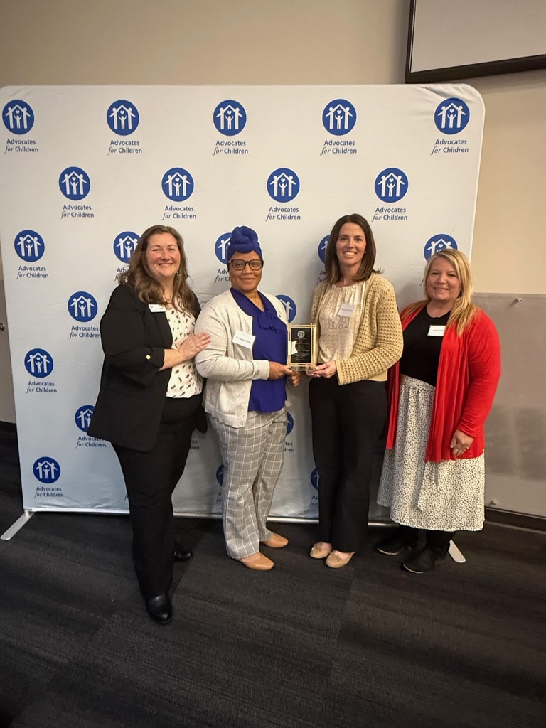 Five women stand together, one holding an award, in front of a blue banner with white logos.