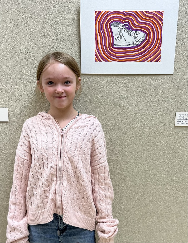 A young girl with blond hair smiles in front of a wall. A framed artwork of a sneaker in a heart shape is mounted above her.