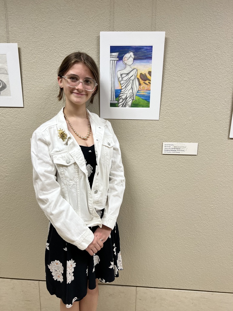 A woman with glasses stands in front of a wall with framed artwork, wearing a white jacket and black dress.