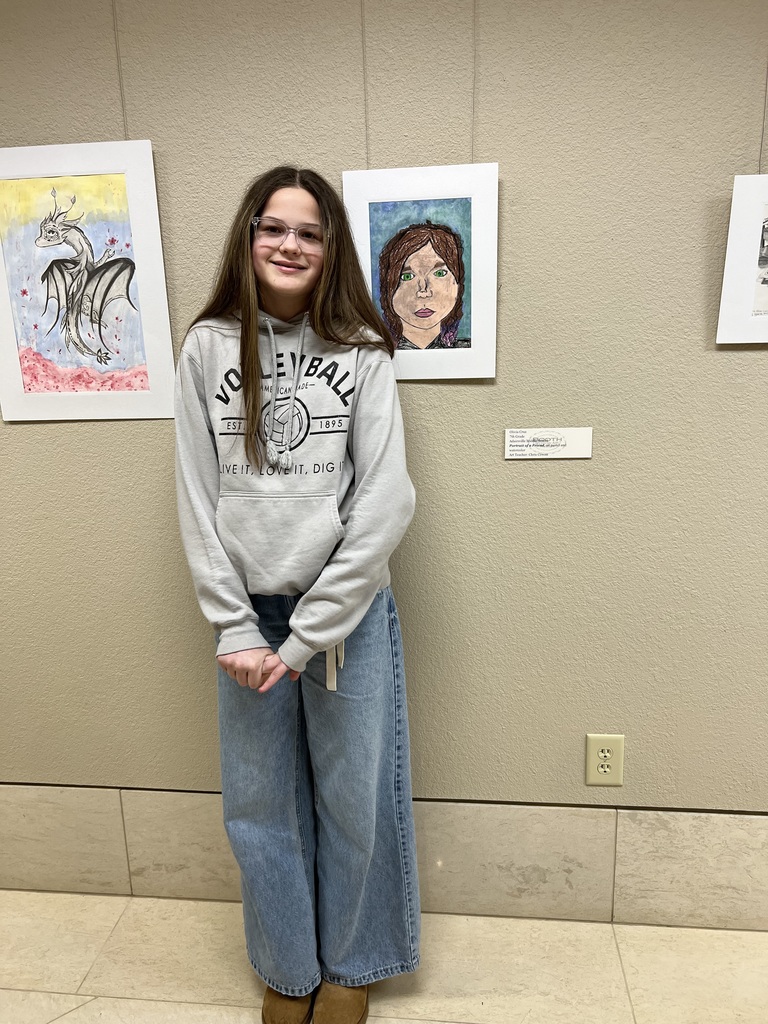A young person stands in front of a wall with framed artwork, wearing a gray sweatshirt and jeans.