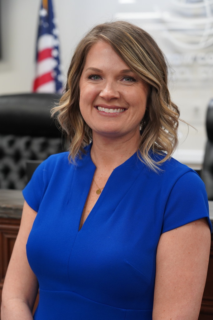 A woman with blonde hair wearing a blue dress sits in an office. An American flag is in the background.