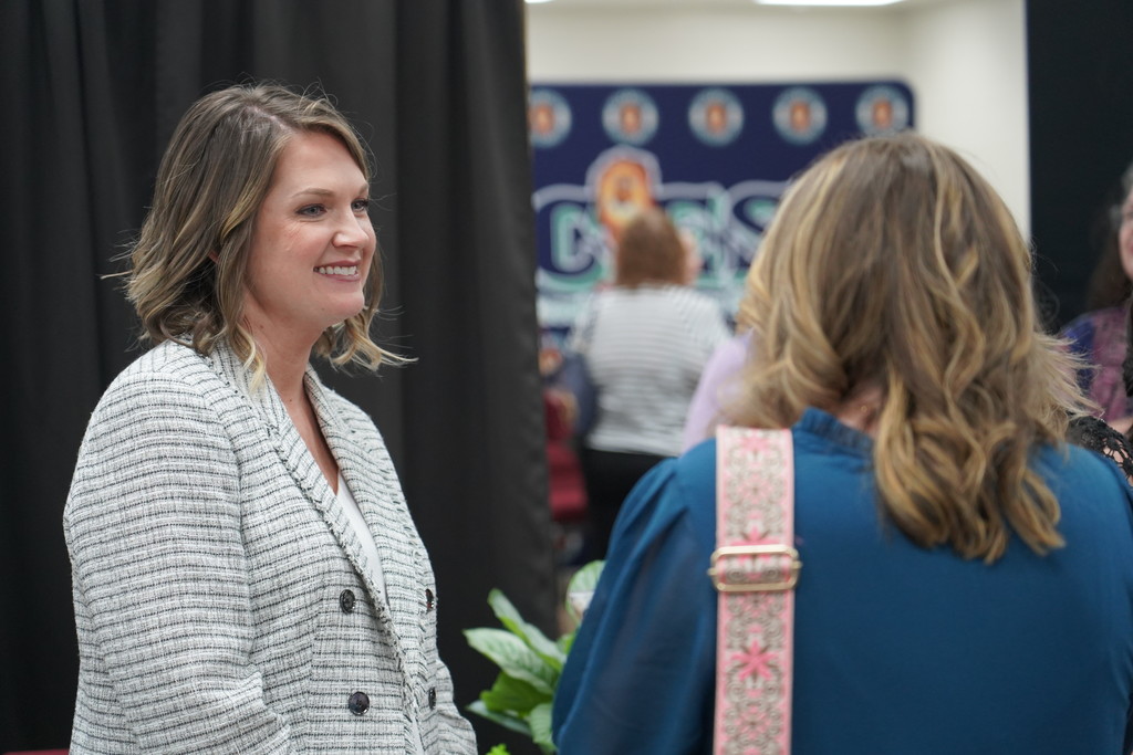 A woman in a gray blazer stands next to another woman with a pink strap on her shoulder. They are talking in a room with a black curtain and a banner.