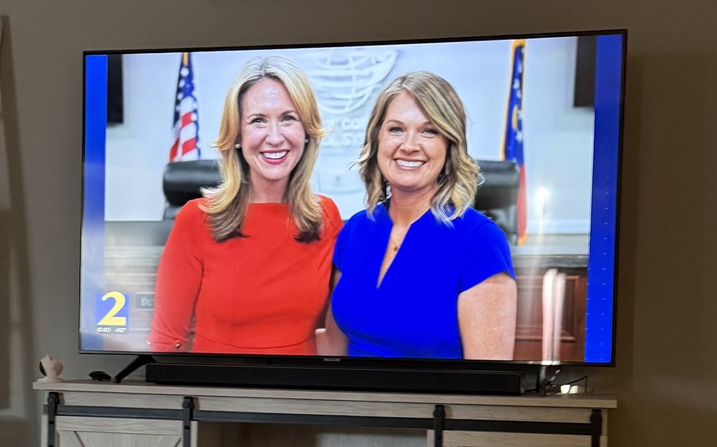 Two women in red and blue tops stand side by side on a TV screen. The screen is mounted on a TV stand.