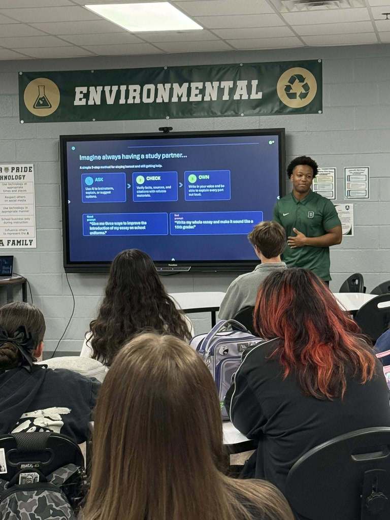 An instructor in a green shirt stands before a monitor displaying a presentation. Students in chairs listen.