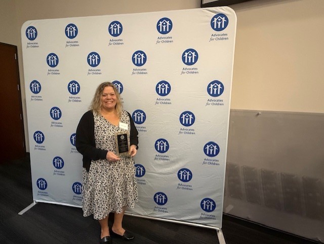 Woman in dress holding award stands in front of a white banner with blue circles and logos.