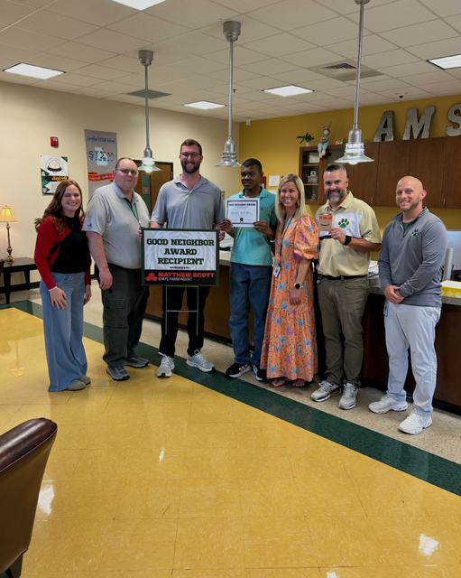Seven people stand in a room holding certificates. They are wearing casual clothing. A yellow floor and lamps are visible.