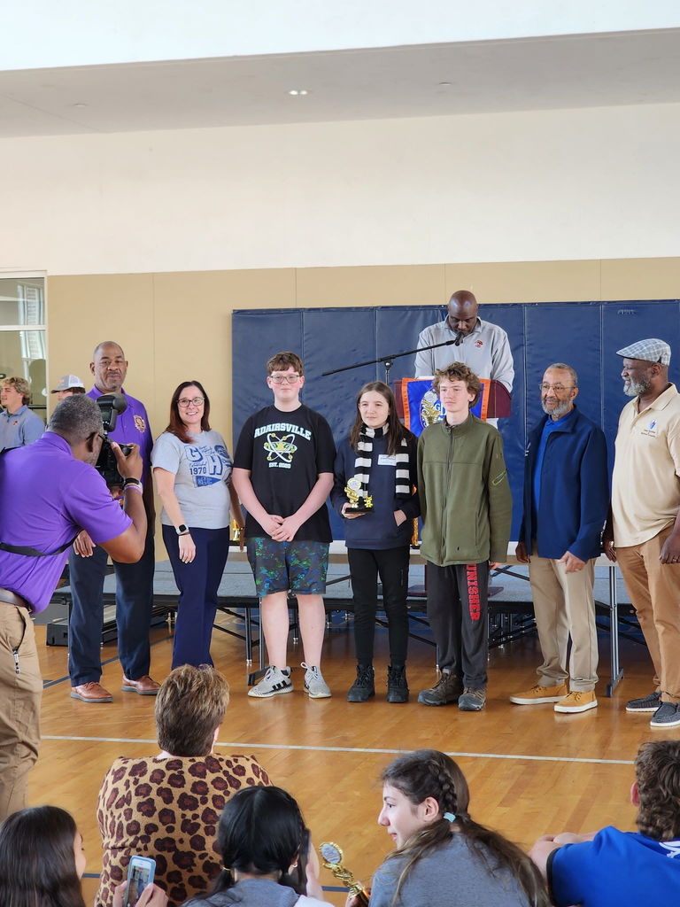 A group of people stand in a circle on a gym floor. One person holds a microphone.
