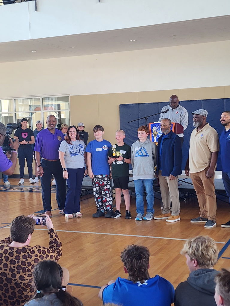 People stand together in a gym. A few hold trophies. A person at a podium speaks into a microphone.