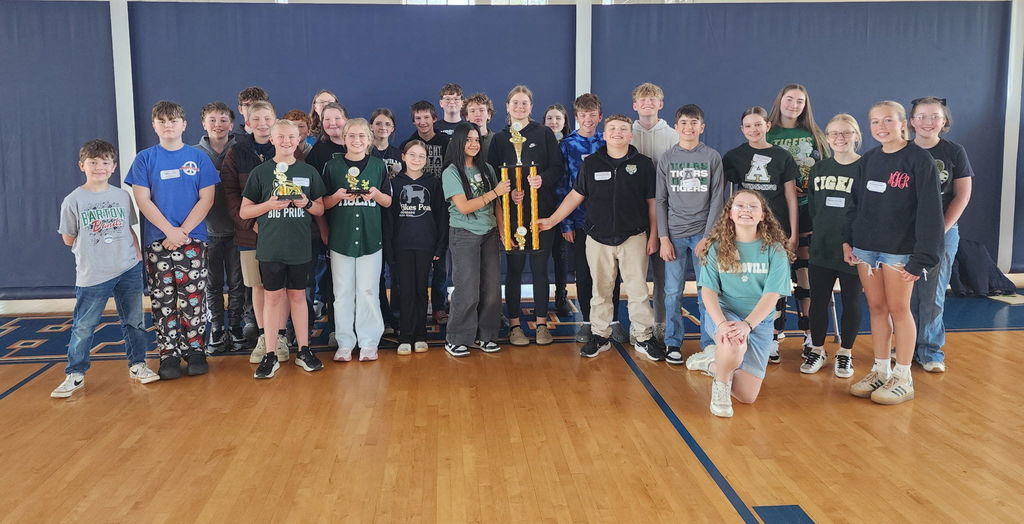 A group of students and staff standing on a gym floor, some holding trophies, with a blue wall in the background.