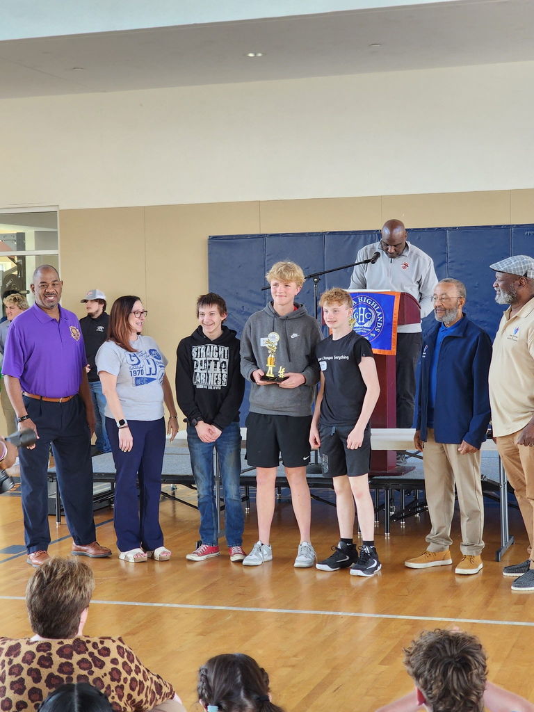 A group of people stands in a gymnasium. A man speaks from a podium, and two boys hold a trophy.