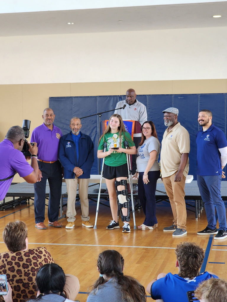 A group of people stands in a gym, with a girl on crutches holding a trophy in front of a podium.