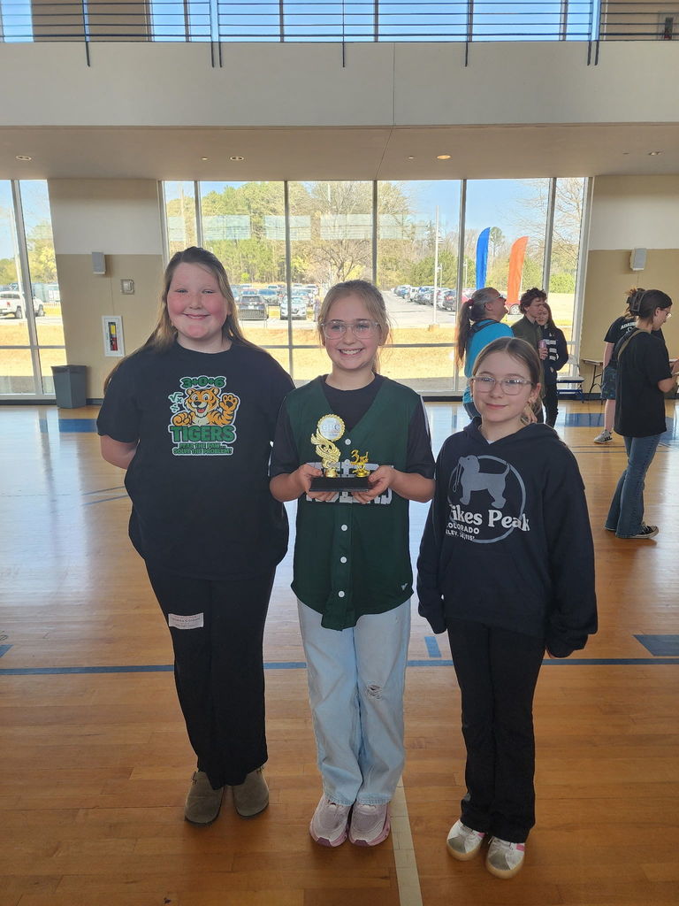 Three girls stand in a school gym, holding a trophy, wearing casual clothes. A wooden floor and glass walls are visible behind them.