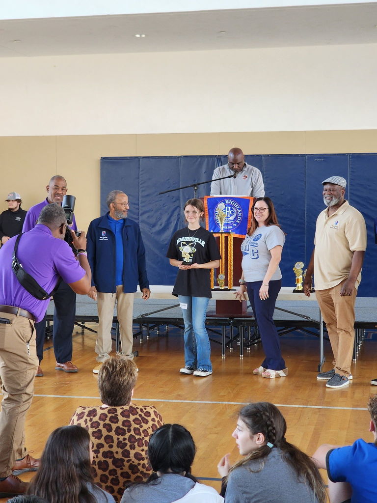 Several adults on a stage, with a woman in front holding an award, while a man speaks behind a podium.