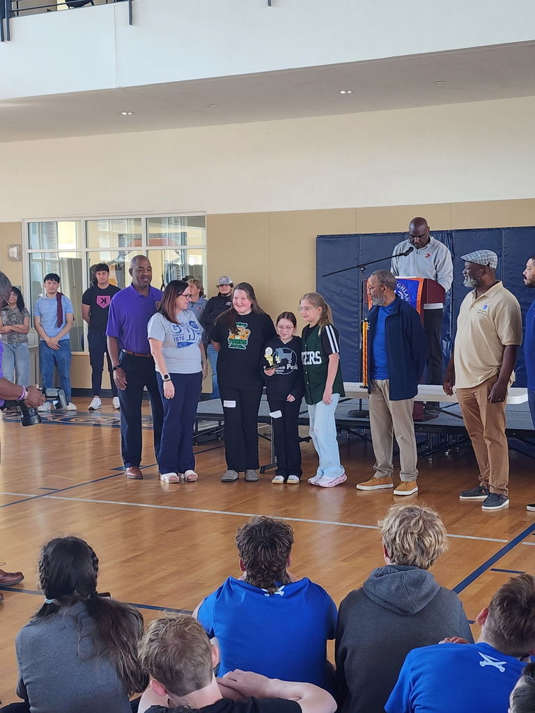 People gather in a gymnasium for an event, with some standing and others sitting. A person speaks from a podium.