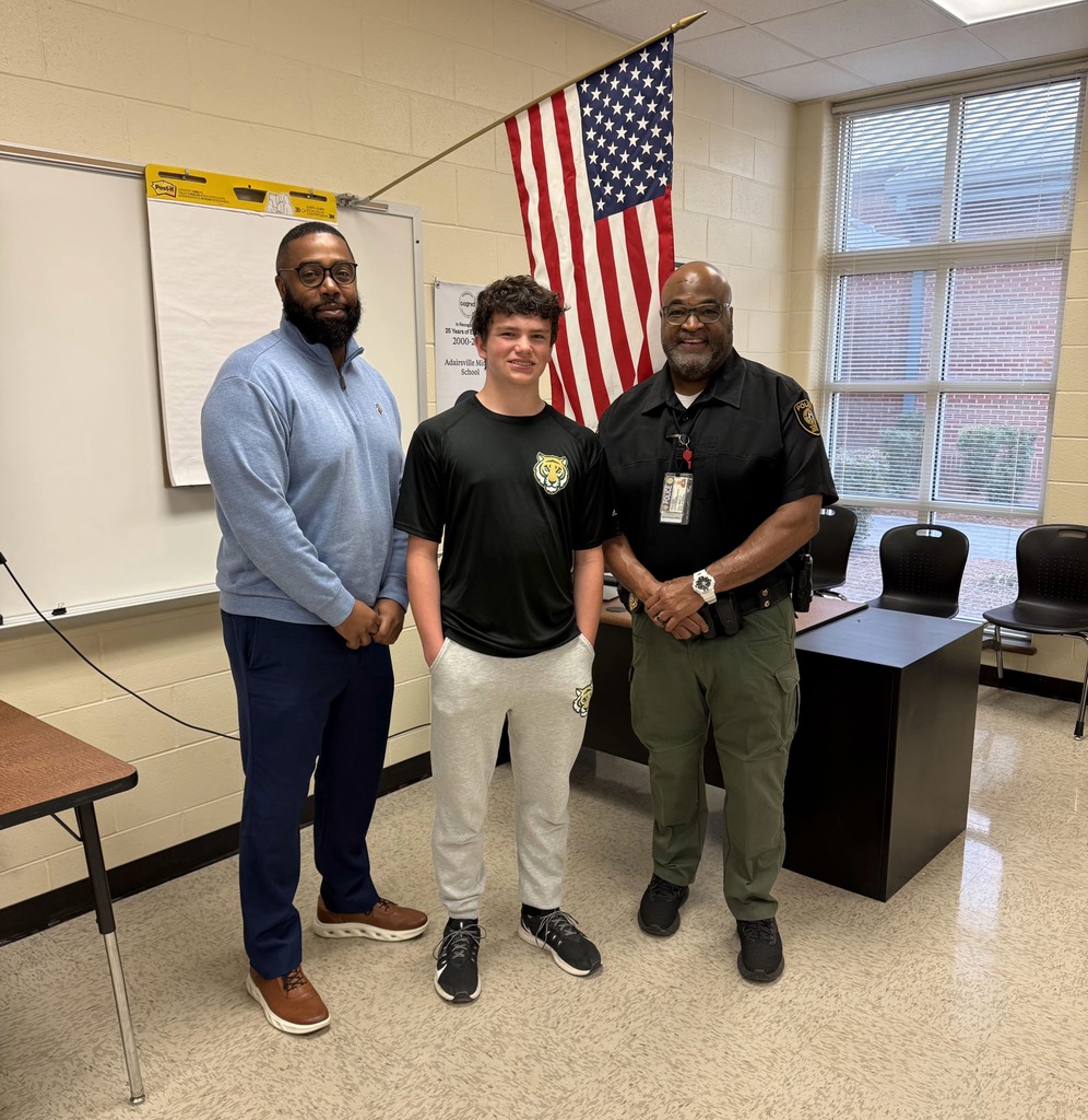 Three men stand together in a classroom. One wears a blue shirt and pants, another a black shirt and pants, and the third a police uniform.
