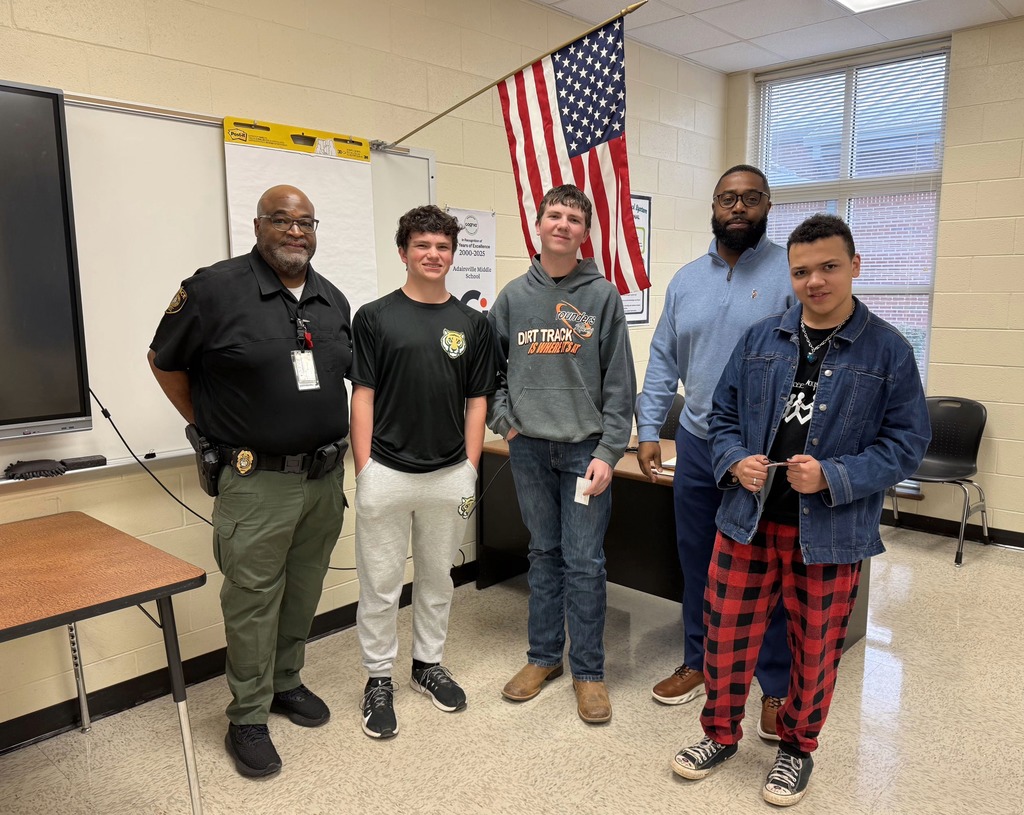 Five people stand in a classroom; two are wearing uniforms. A man in a police uniform holds a belt.