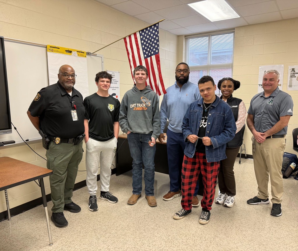 Several people stand in a classroom. A man in a police uniform stands in front of them.