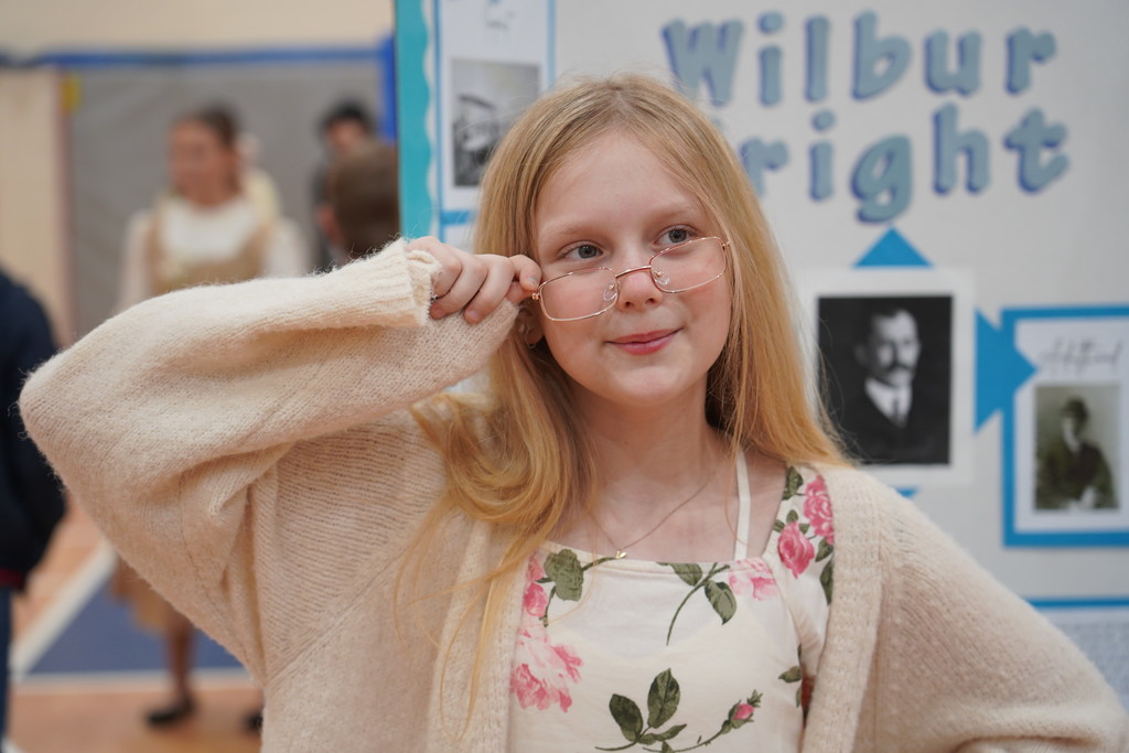 A young girl with blonde hair holds up glasses, smiling, in front of a bulletin board.