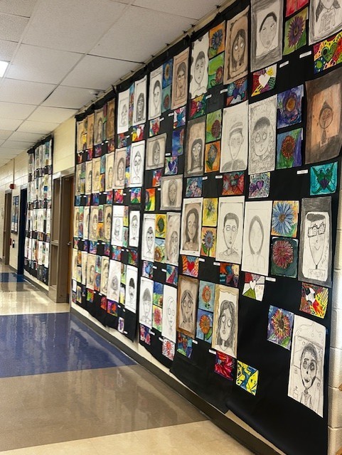 Hallway with a wall covered in various colorful drawings, from portraits to abstract art, in a school setting.