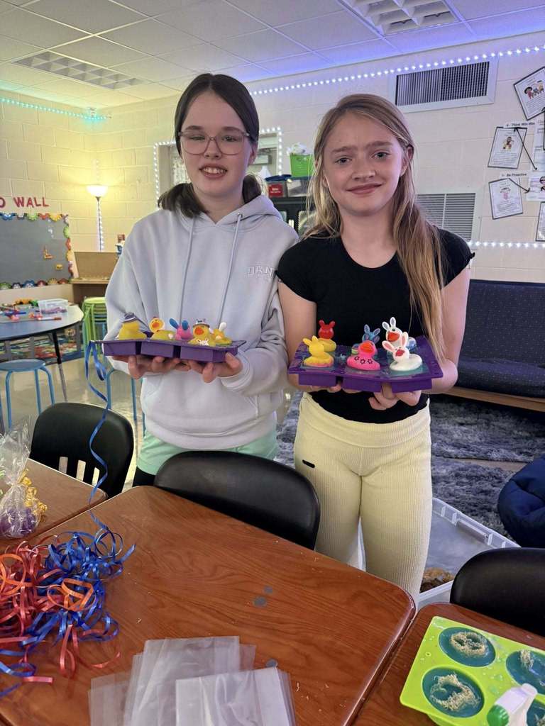 Two girls hold trays of cupcakes in a classroom setting. One girl wears a white hoodie, the other a black shirt and yellow pants.