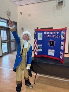 A child dressed in historical costume points to a display with "Alexander Hamilton" written on it.