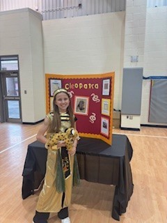 A young girl in an ancient costume stands next to a table with a display about Cleopatra.
