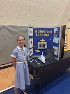 A girl in a blue dress stands in front of a display about Katherine Johnson, featuring photos and a small model.