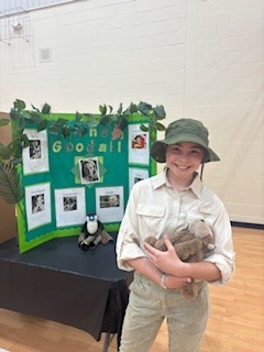 Person in safari hat holding stuffed animal in front of green display with photos.