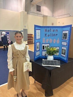 A young girl in a historical dress stands beside a table. The table has a book, flowers, and a sign with the name Helen Keller.