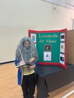 A person in a long gray wig and blue robe stands next to a display board.