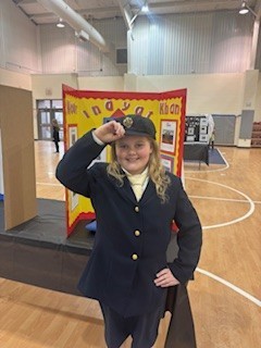 A young girl in a navy blue uniform, standing indoors, with a podium and a colorful sign behind her.
