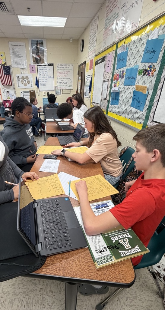 Five students and an adult sit around a table with laptops, papers, and a book. The room has white walls, bulletin boards, and a flag.