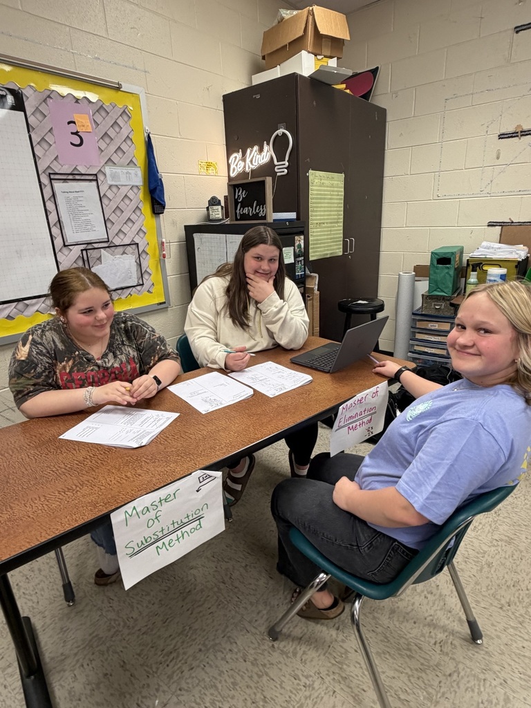 Three individuals seated at a table in a classroom, with papers and a laptop, one wearing a blue shirt, and another wearing a white shirt.