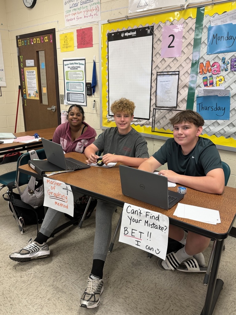 Three students sit at a table with laptops, one holding a sign that reads, "Can't Find Your Mistake?" Behind them is a classroom wall with papers and a bulletin board.
