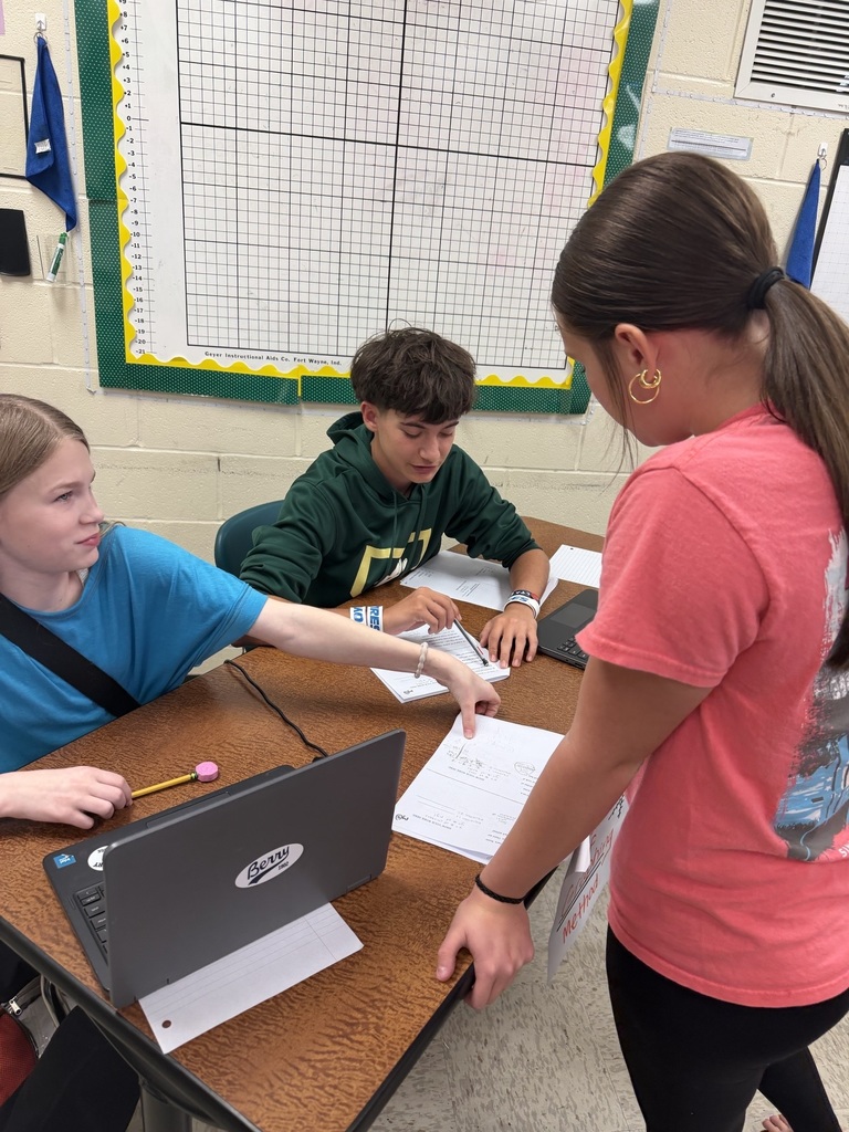 Three students seated around a desk, one holding a pencil and papers, another holding a laptop, and the third holding a piece of paper.