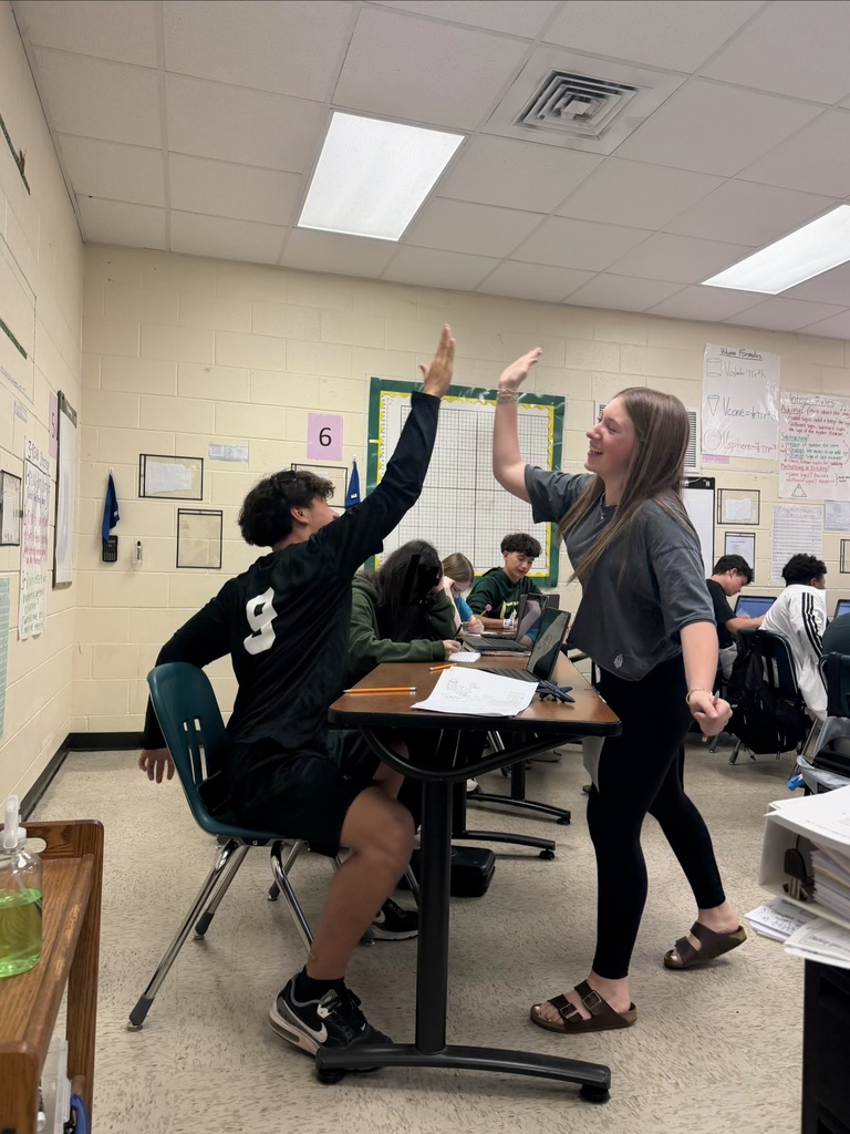 Two students, one seated and one standing, play rock-paper-scissors near a desk in a classroom.