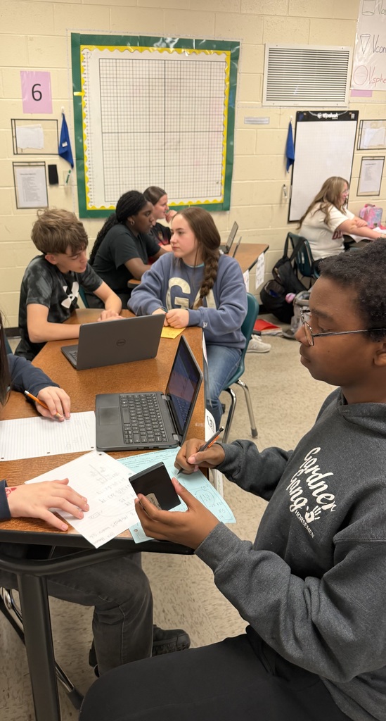 A classroom with yellow walls, where several students work at tables. One student is writing on a piece of paper while another holds a cell phone.