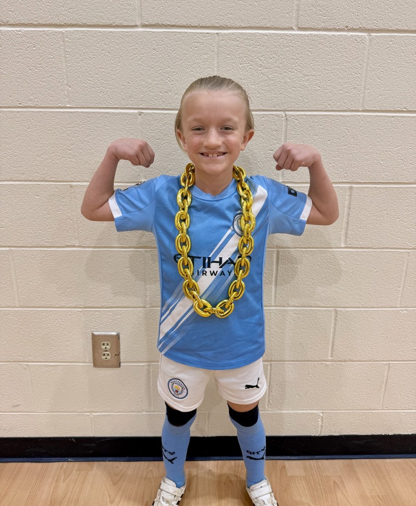 A young person in a blue soccer uniform with a gold chain stands in a room with a white wall.