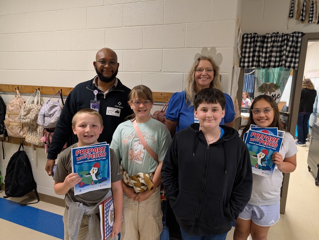 A group of students and two adults pose for a photo in a classroom with bags hanging on the wall.
