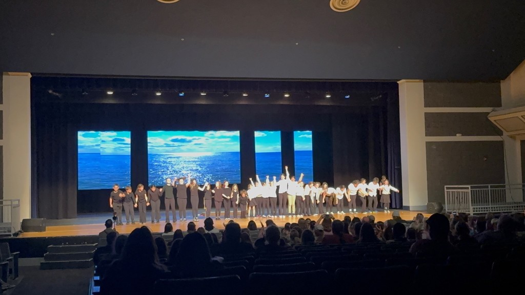 Performers on stage with large screens behind them, ocean view projected. Audience seated in front.