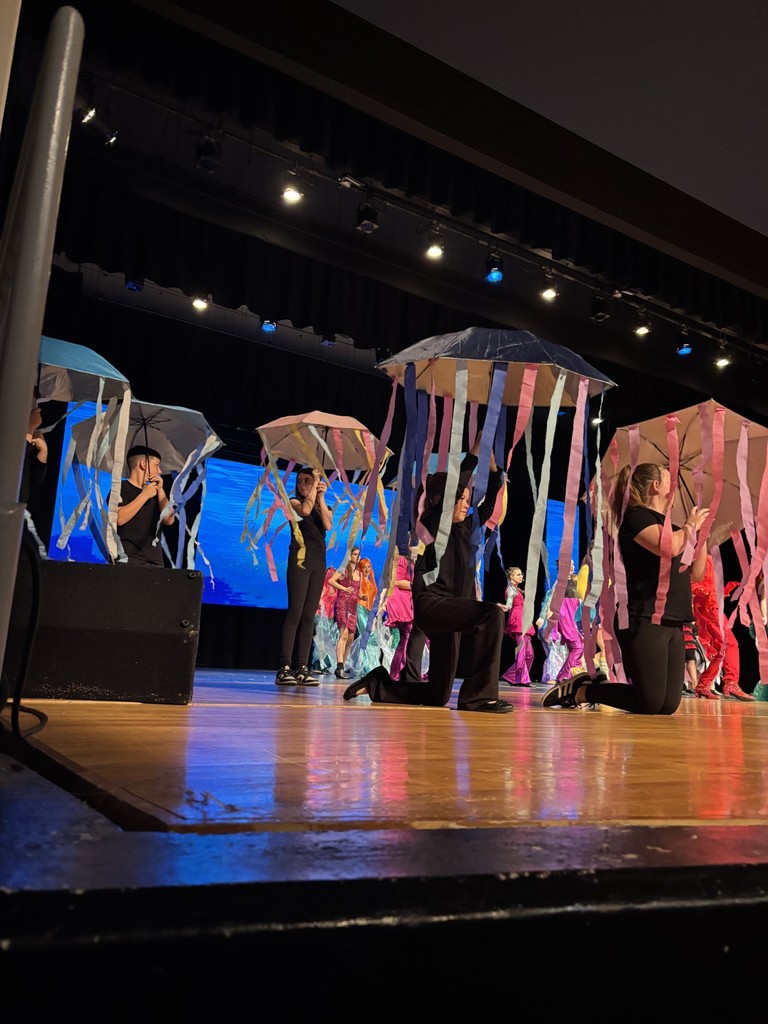 People on a stage perform a dance with umbrellas; multicolored ribbons float from the umbrellas.