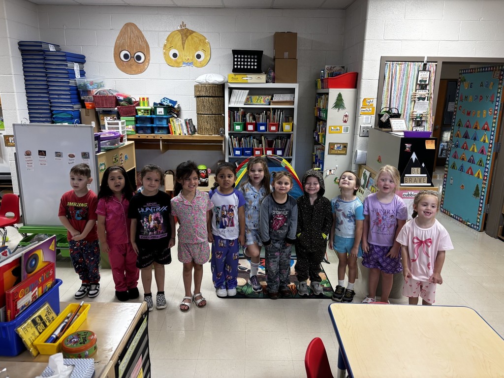 Children in matching pajamas stand together in a classroom with shelves, toys, and a refrigerator.