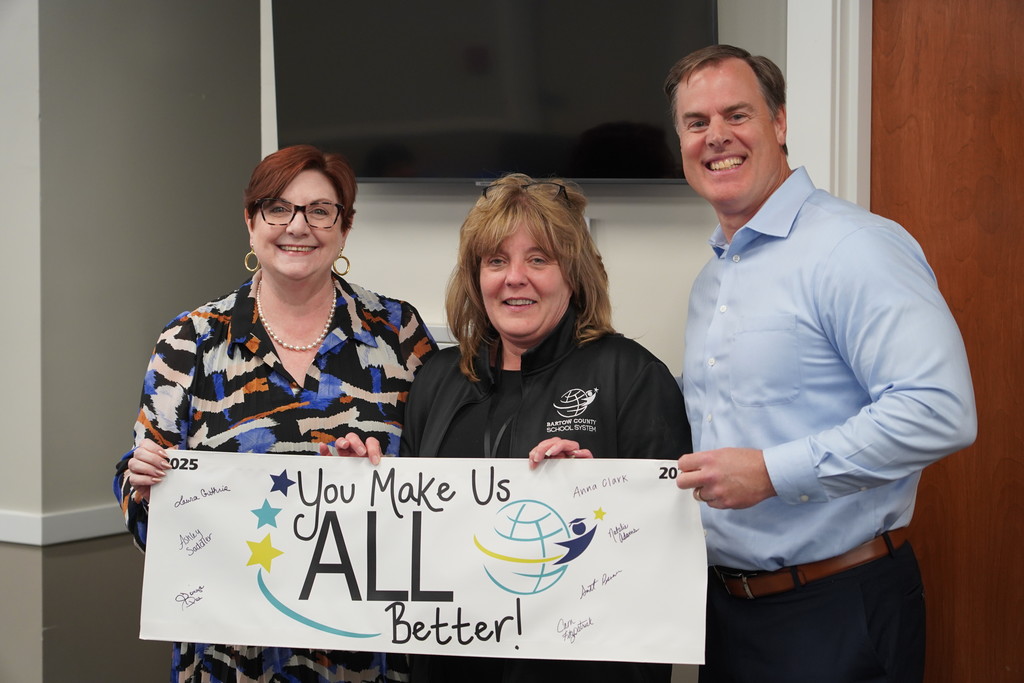 Three people stand smiling, holding a sign with the words "You Make Us All Better!" and an image of a globe.