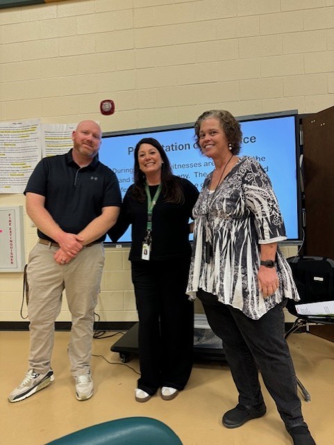 Three adults stand together before a projector screen displaying text in a classroom setting.