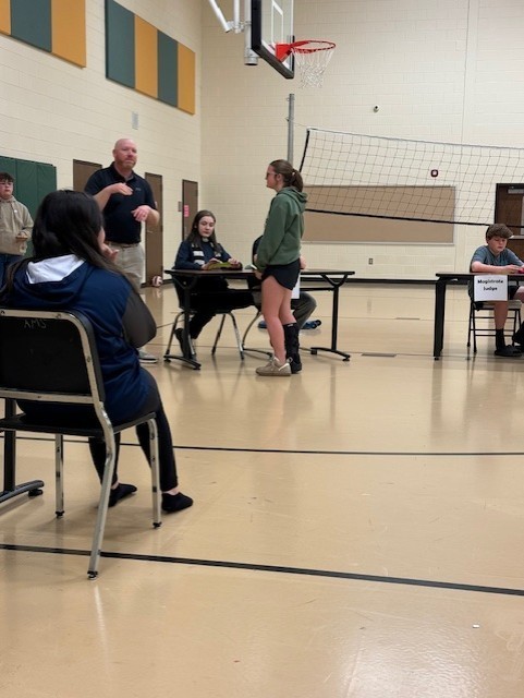 A basketball court with a volleyball net. Several people sit at tables, one person stands, and another is seated in a chair.