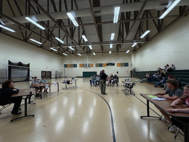 Gymnasium with multiple tables. Some people sit at tables while others sit in bleachers. People in the middle of the gym stand and listen.