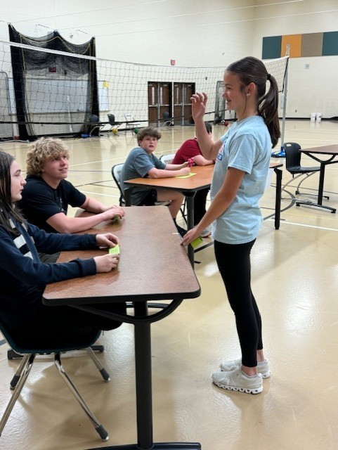 A girl in a blue shirt stands in front of seated teens at a table in a gym.