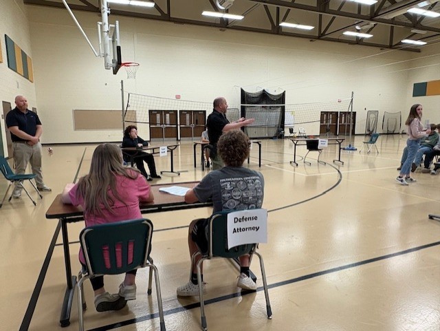 Several people sit at tables inside a gymnasium. A man stands at the head of the tables, facing a group.
